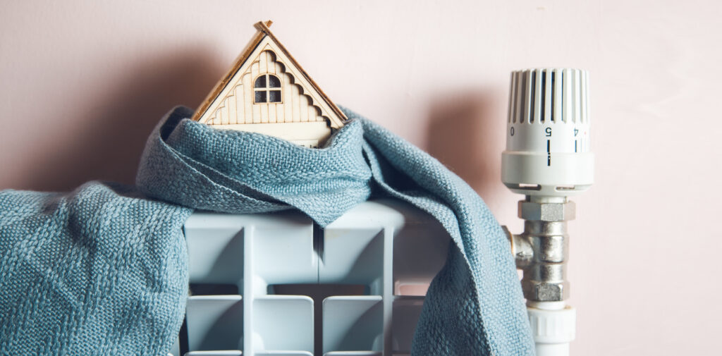 Foto für Blog Beitrag ELHADO Im Winter richtig heizen. Ein Modell eines Hauses steht auf einem Heizkörper und ist in einen Schal eingewickelt