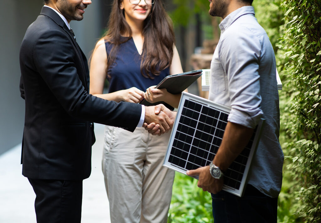 Energieberater, der das Modell einer Photovoltaikanlage unterm Arm hält, schüttelt die Hand eines betrieblichen Mitarbeiters. Neben ihnen steht eine Mitarbeiterin, die ein Tablet in der Hand hat.