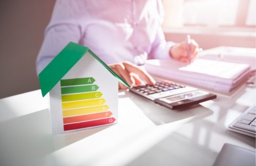 Bild zu Individueller Sanierungsfahrplan. Person sitzt am Schreibtisch und gibt Zahlen in Taschenrechner ein, gleichzeitig macht sie Notizen. Vor ihr steht ein Häusermodell, auf dessen Vorderseite sich die Energieskala befindet.