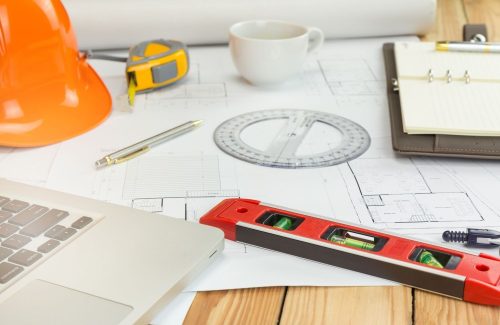 Ausschnittsfoto von Schreibtisch mit Macbook, Bauhelm, Maßband, Kugelschreiber, Lineal, Kaffeetasse, Terminplaner, Wasserwaage und Grundrissen einer Immobilie - Foto für Baubegleitung bei ELHADO Engineering