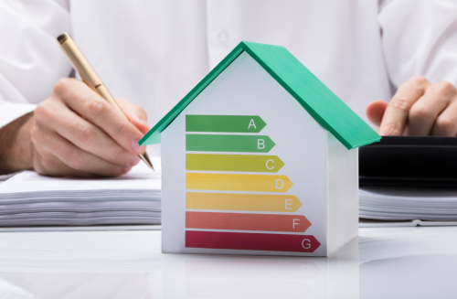 Foto für Initialberatung. Energieberatung. Person schreibt Notizen. Vor ihr auf dem Schreibtisch steht ein Hausmodell, auf dessen Vorderseite eine Energieeffizienz-Skala abgebildet ist.